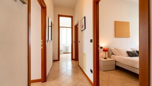 Un couloir d’hôtel avec des portes en bois, un sol carrelé, un lit avec une table de chevet, une lampe et un radiateur.