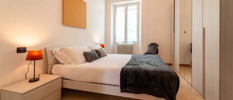 Un dormitorio con cama, mesitas de noche, lámpara, radiador y ventana.