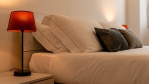 Un lit bien fait, avec des oreillers blancs et foncés, une lampe rouge sur une table de chevet et une table de nuit en bois.