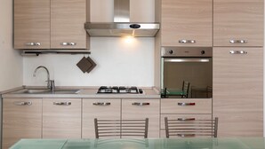 Une cuisine moderne avec des meubles en bois clair, des appareils électroménagers en acier inoxydable, une cuisinière à gaz et une table à manger en verre, assortie de chaises.