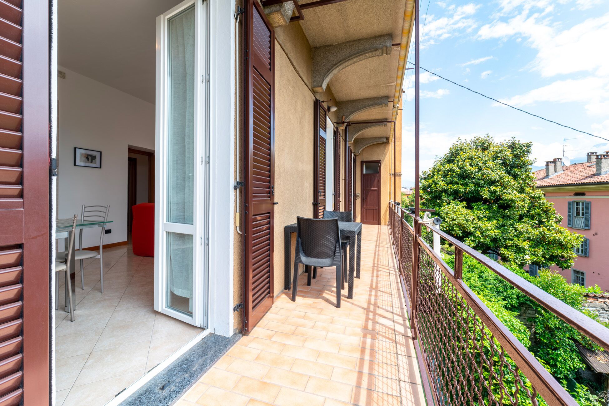 A balcony with a table and chairs, a red ottoman, and a view of a tree and buildings.