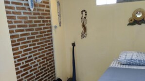 Un dormitorio con pared de ladrillo, una cama con colcha azul, un ventilador de pie, un cesto de basura y un adorno colgante en la pared.