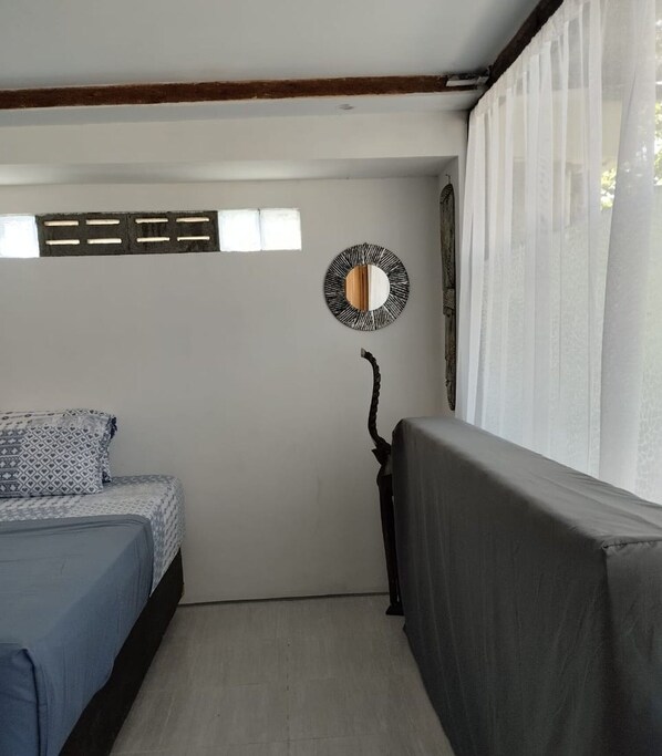 A small, compact room with a bed, a chair, a wall-mounted mirror, and a window with curtains. - Adem Huis Samen (Sumbermulyo)