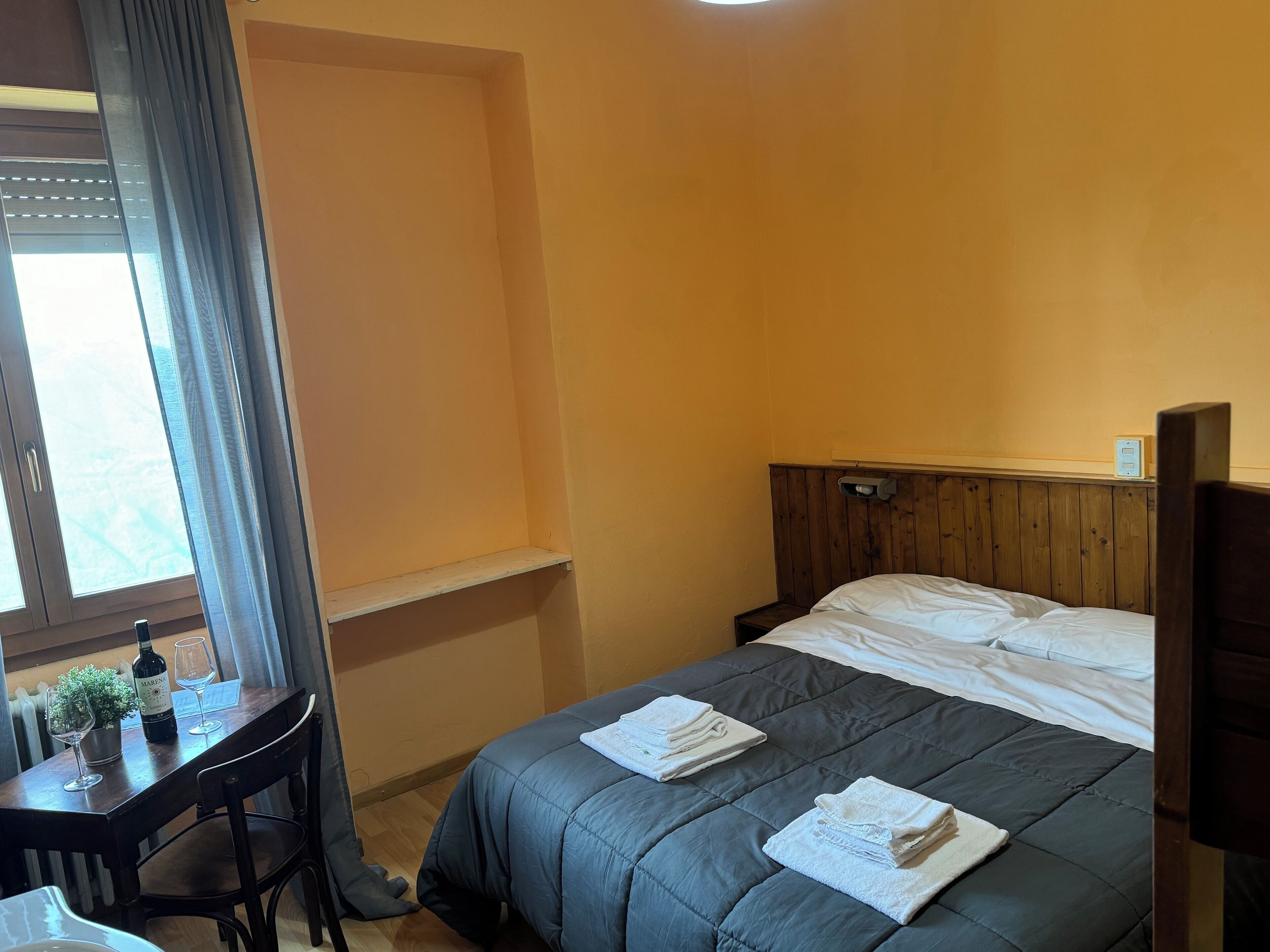 A hotel room with a bed, a small table with a bottle and glasses, a window with curtains, and a wooden headboard.