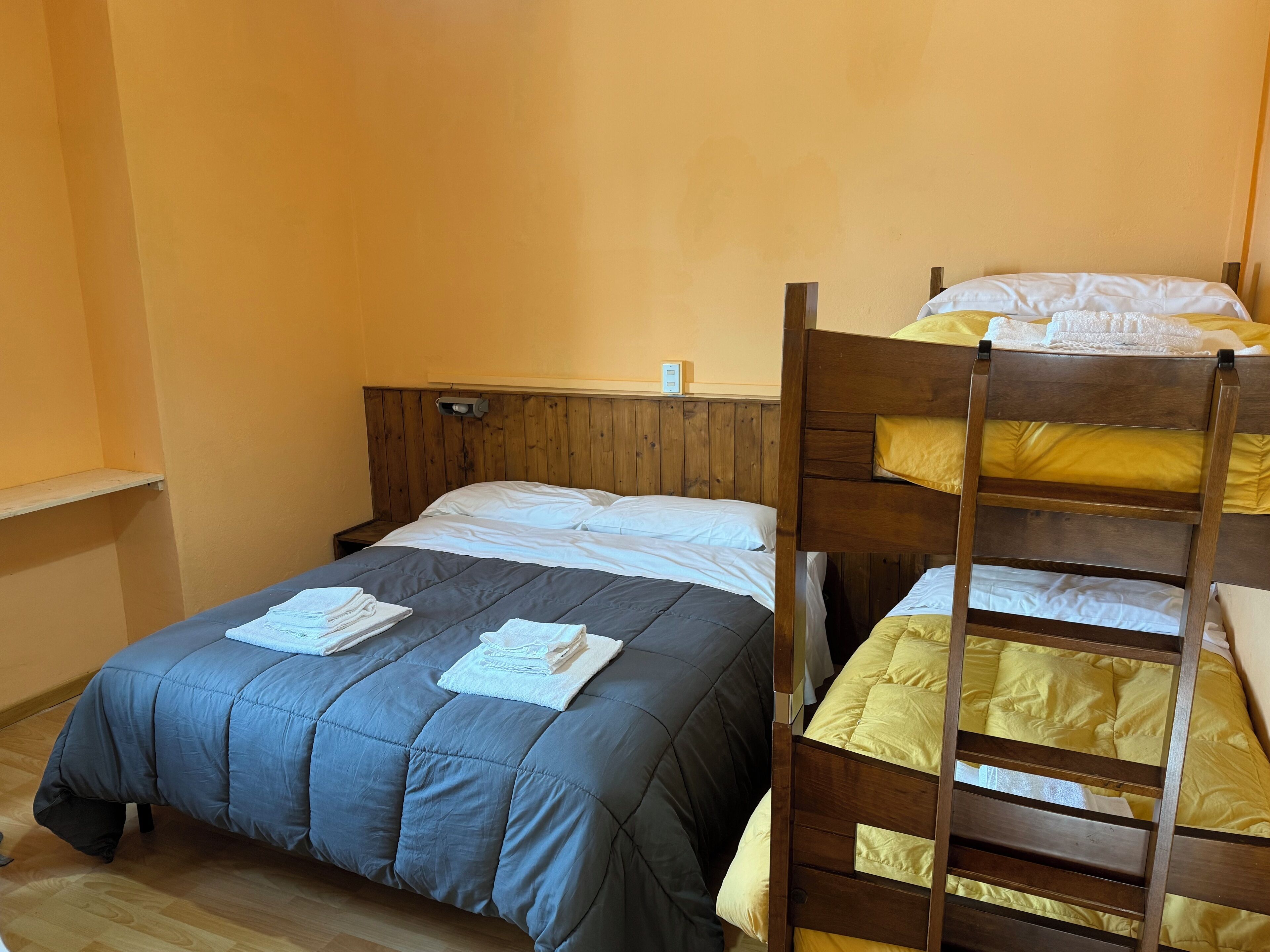 A room with a bunk bed, a single bed, wooden headboard, a bedside table, and a wall-mounted light.
