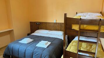 A room with a bunk bed, a single bed, wooden headboard, a bedside table, and a wall-mounted light.
