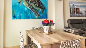 A dining area with a wooden table, white chairs, a painting of a sea turtle, and a wall-mounted surfboard art piece. - Detinsa by Interhome (Sant Jordi)