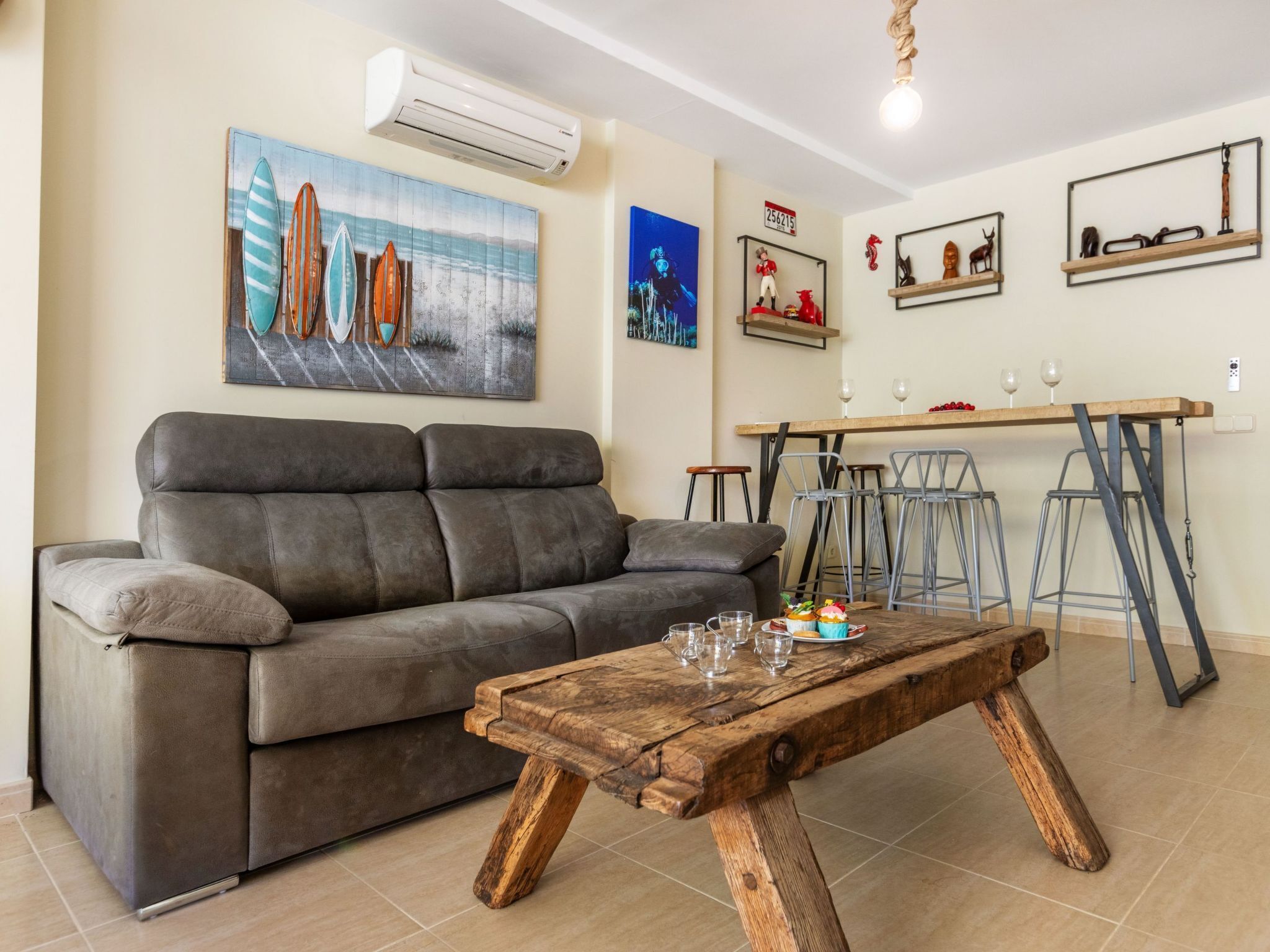 A living room with a grey sofa, a wooden coffee table with glasses and a cupcake, a bar area with stools, and wall art including surfboards and a cityscape.