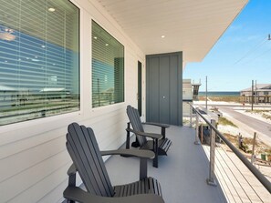 Un balcón con sillas Adirondack, vista a la playa y un edificio con grandes ventanales.