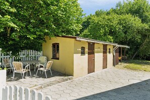 Ein kleines, gelbes Gebäude mit einer Tür, einem Fenster und einem weißen Zaun, sowie ein Tisch mit Stühlen davor.
