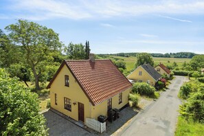 Une maison jaune avec un toit rouge, une allée en gravier et une clôture blanche faite de piquets.