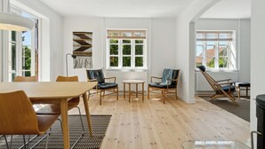 A spacious room with a wooden dining table, two chairs, a small round table, and a sofa. There are large windows with blinds and a view of greenery outside.