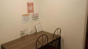 Uma pequena área de refeições com uma mesa de madeira e quatro cadeiras. Há placas na parede, incluindo um aviso sobre pertences perdidos e proibição de fumar ou usar vaporizadores.