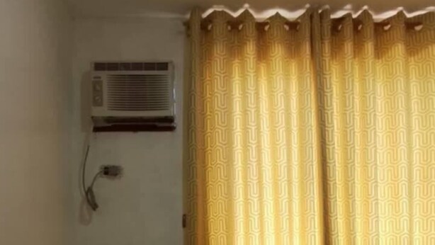 Quarto de hotel com cama, travesseiro em formato de coração vermelho, ar condicionado fixado na parede e cortina com padrão geométrico.