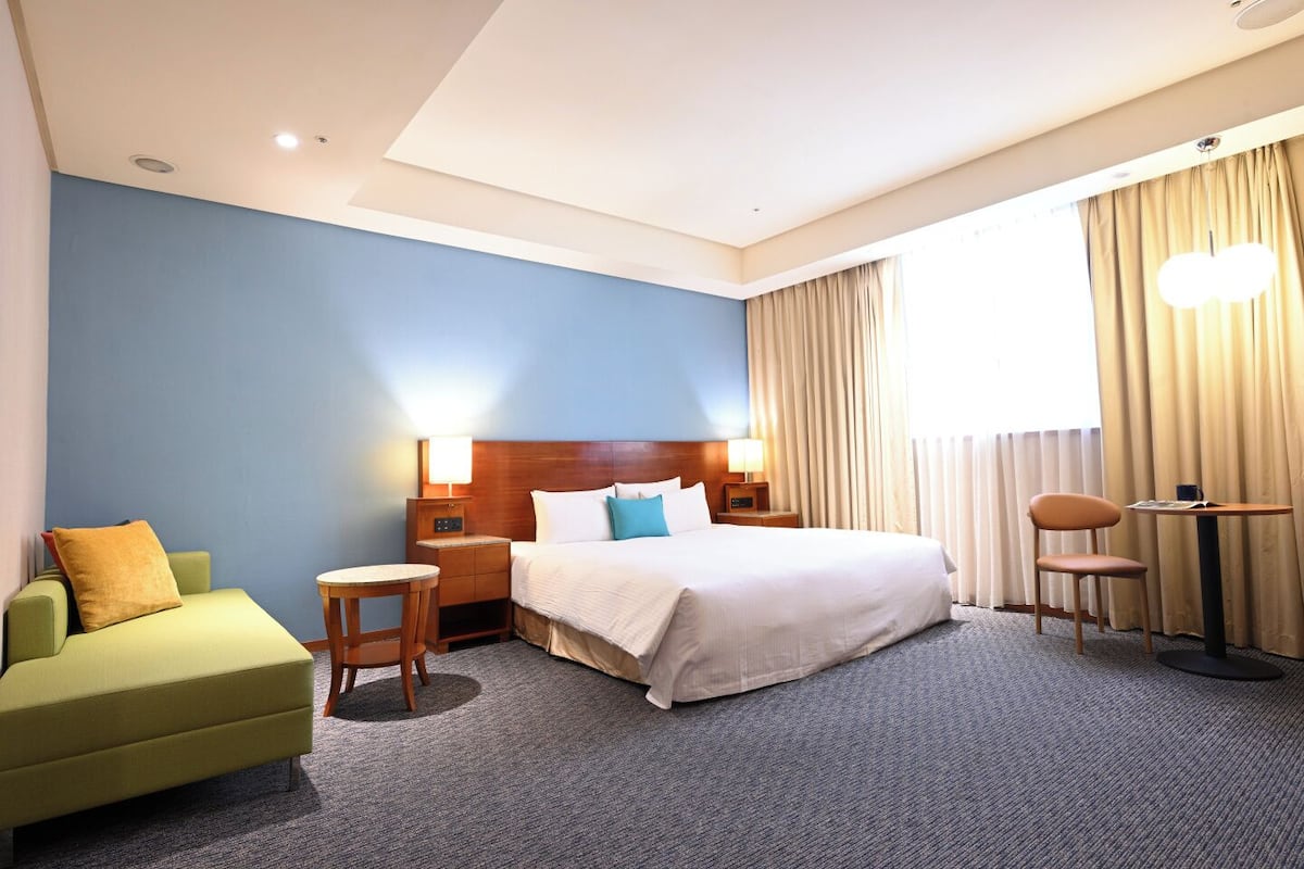 A hotel room with a bed, a green armchair, a small table, and a chair.