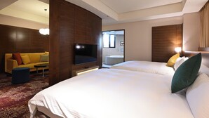Habitació d'hotel amb dos llits, un sofà, una televisió i un bany visible a través d'una porta oberta.