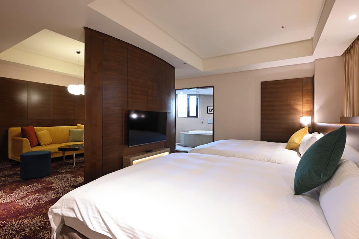 A hotel room with two beds, a sofa, a TV, and a bathroom visible through an open door.