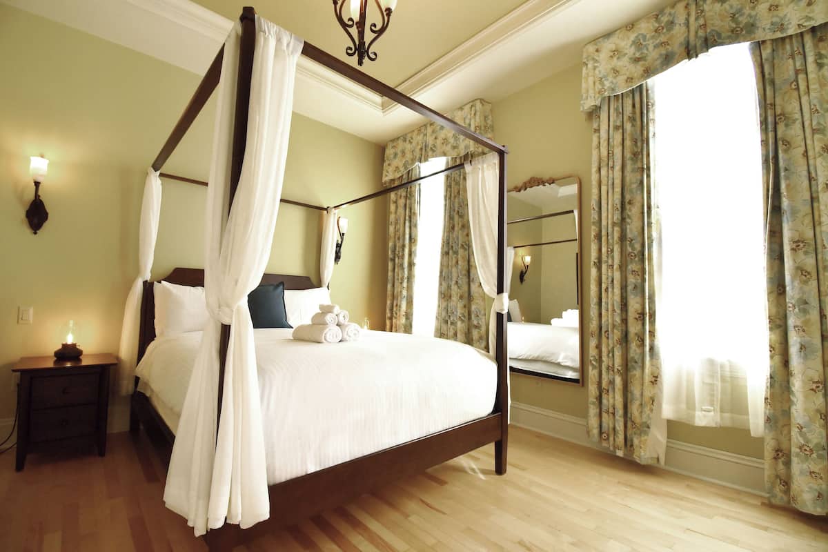 A four-poster bed with white linens, a bedside table with a lamp, and a large mirror on the wall.