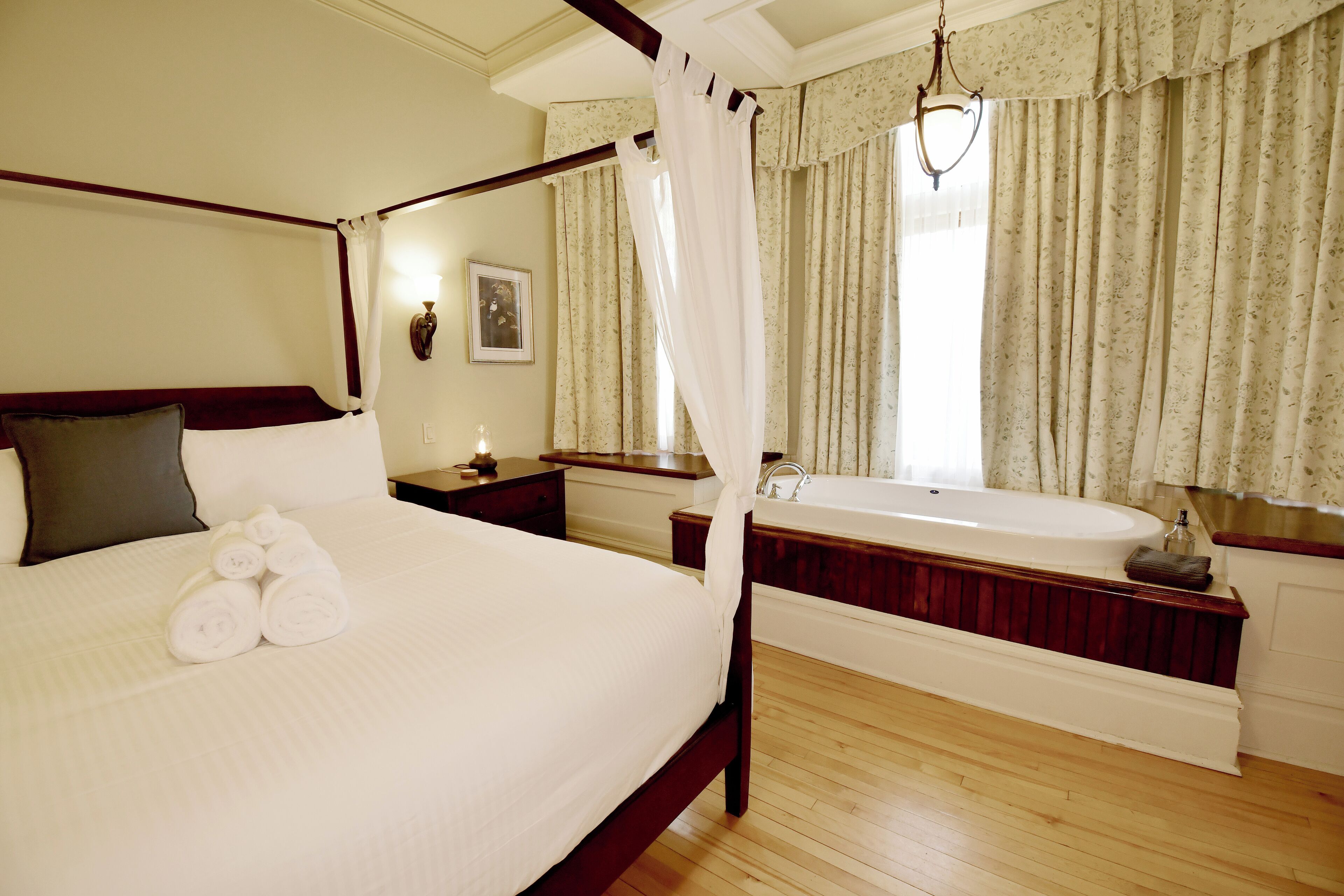 Una cama con dosel, toallas enrolladas, cabecera de madera, mesita de noche, espejo, ventana grande con cortinas y suelo de madera.