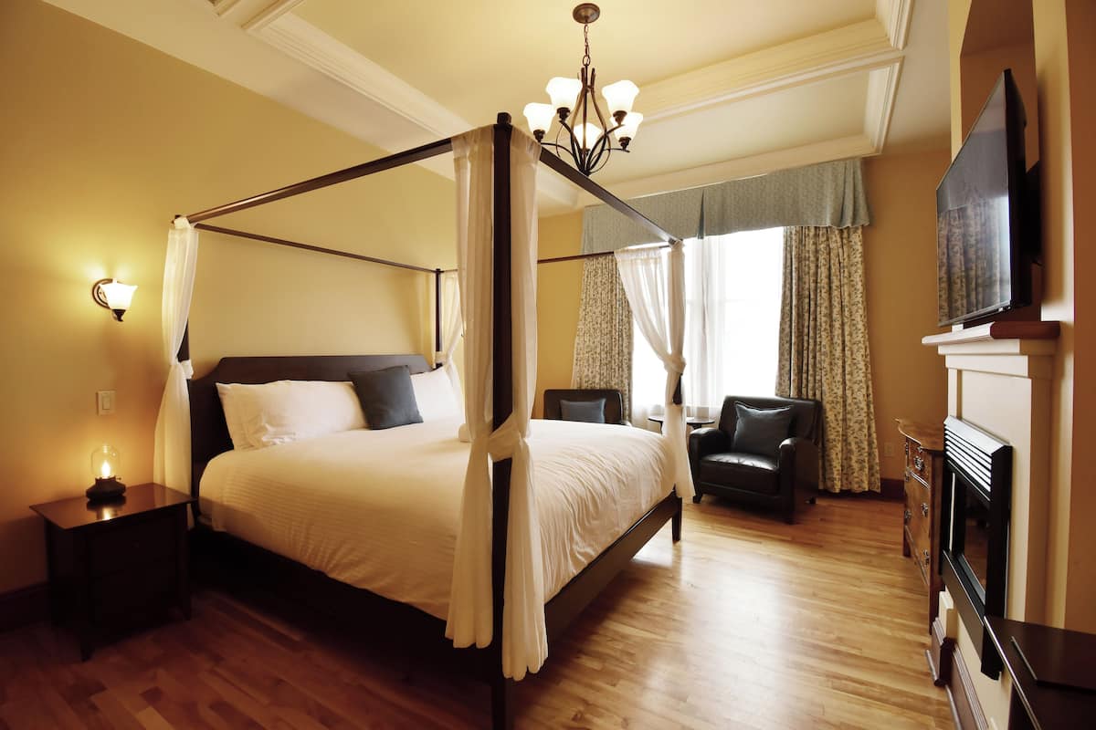 A four-poster bed with a canopy, a television mounted on the wall, a fireplace, and a seating area with a chair and table.