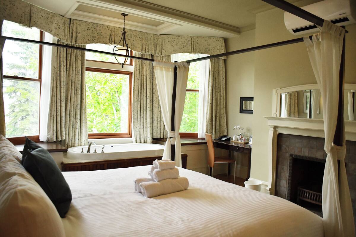 A hotel room with a large bed, a bathroom with a bathtub, a fireplace, and curtains on the windows.