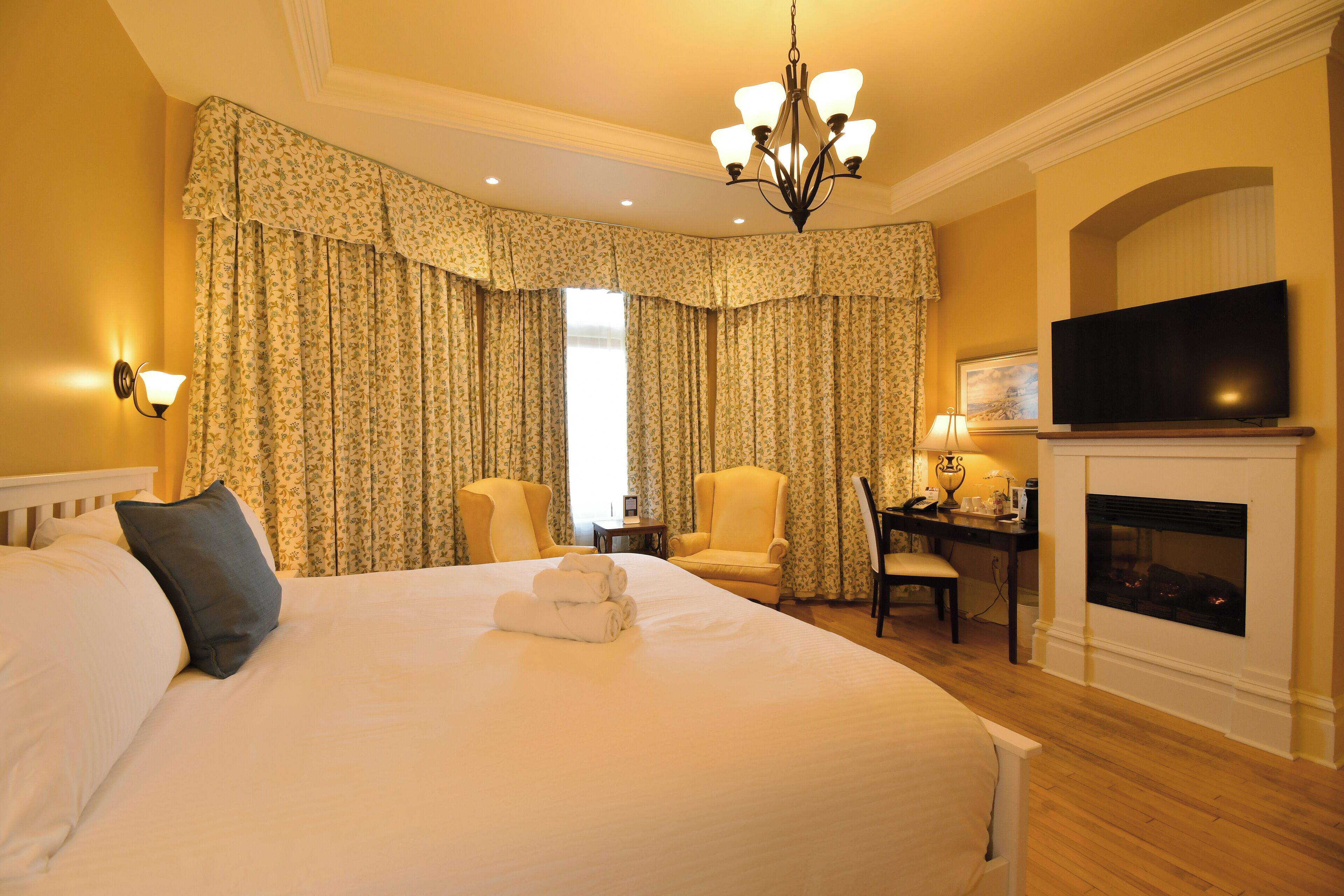 A hotel room with a large bed, two armchairs, a desk with a lamp, a television, and a fireplace.