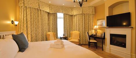 A hotel room with a large bed, two armchairs, a desk with a lamp, a television, and a fireplace.