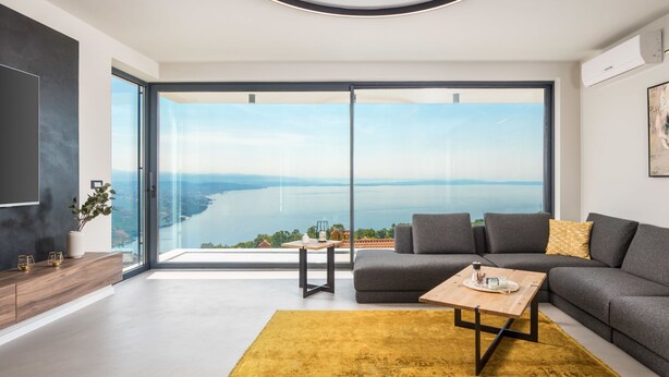 A modern living room with a large window offering a view of a lake and distant mountains. There is a gray sofa, a wooden coffee table, and a yellow rug. - Opatija Sky by Interhome (Opatija/Bregi)