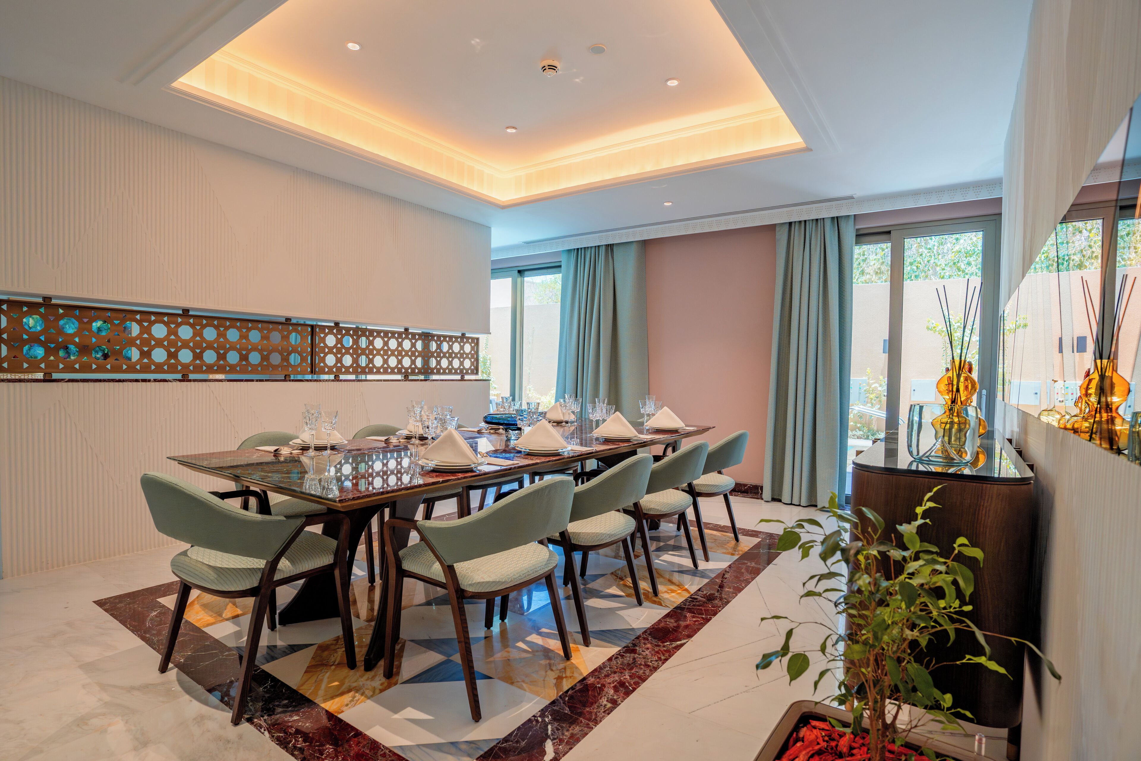 Un coin repas avec une longue table dressée pour six personnes, entourée de chaises, agrémenté d’un panneau décoratif mural et d’une plante en pot.