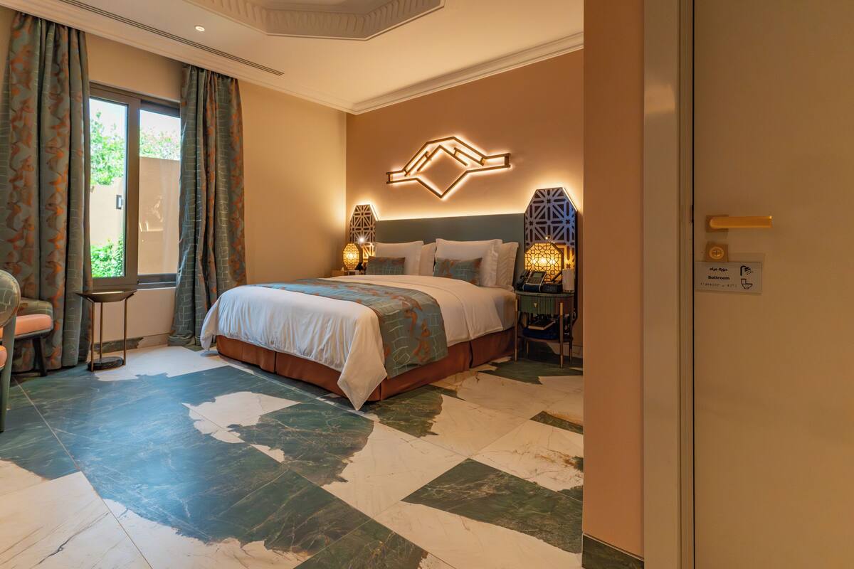 A hotel room with a bed, a nightstand, a lamp, and a view of the outdoors.