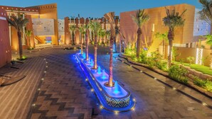 Un patio de hotel bien iluminado con fuente de agua, palmeras y arquitectura moderna.