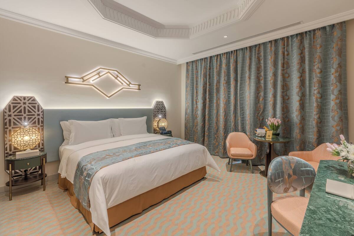 A hotel room with a large bed, two bedside tables with lamps, a desk with a chair, and a large window with curtains.