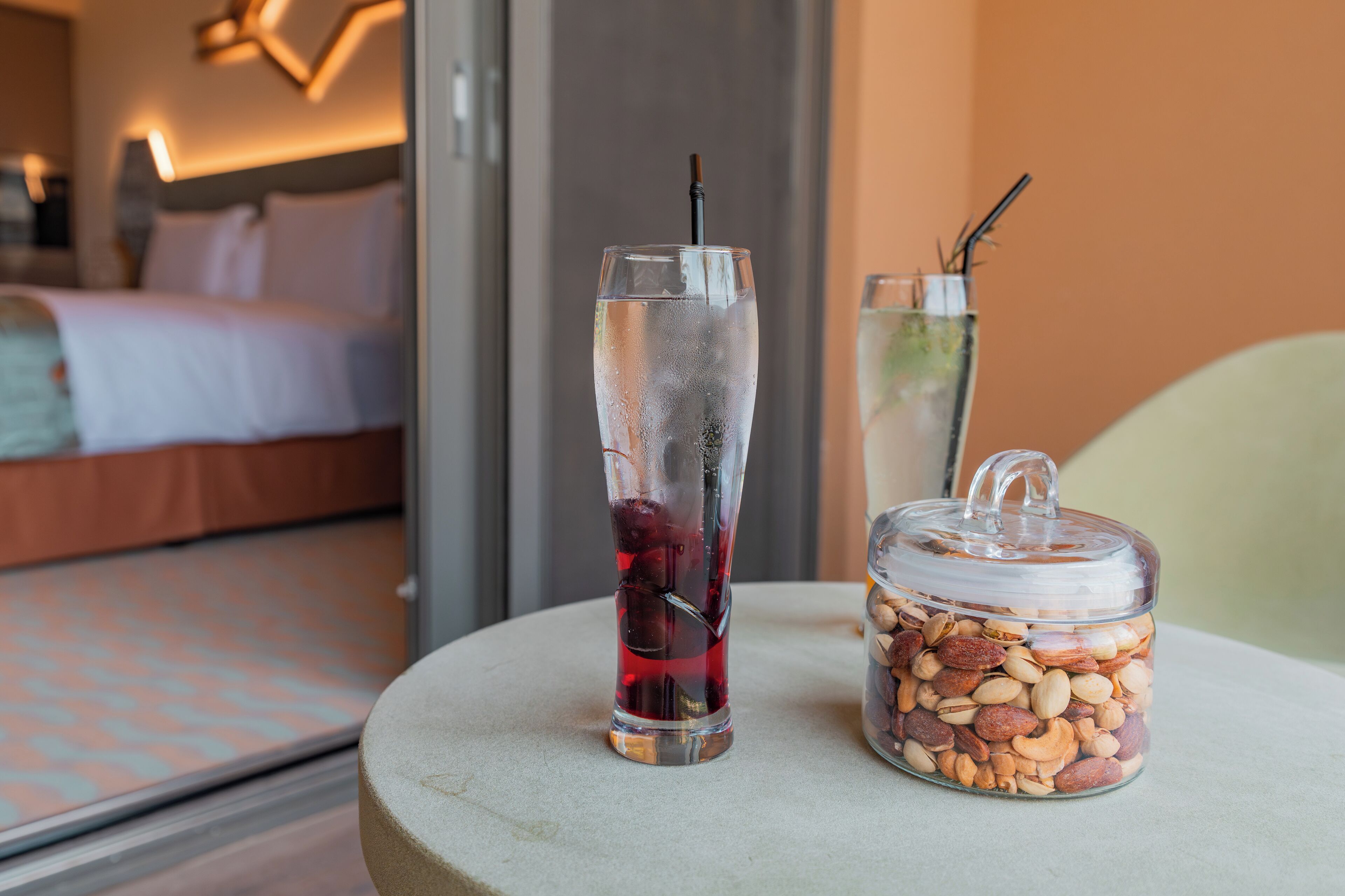 Een glas met ijskoud drankje en een rietje, een potje notenmix, en een bed op de achtergrond.