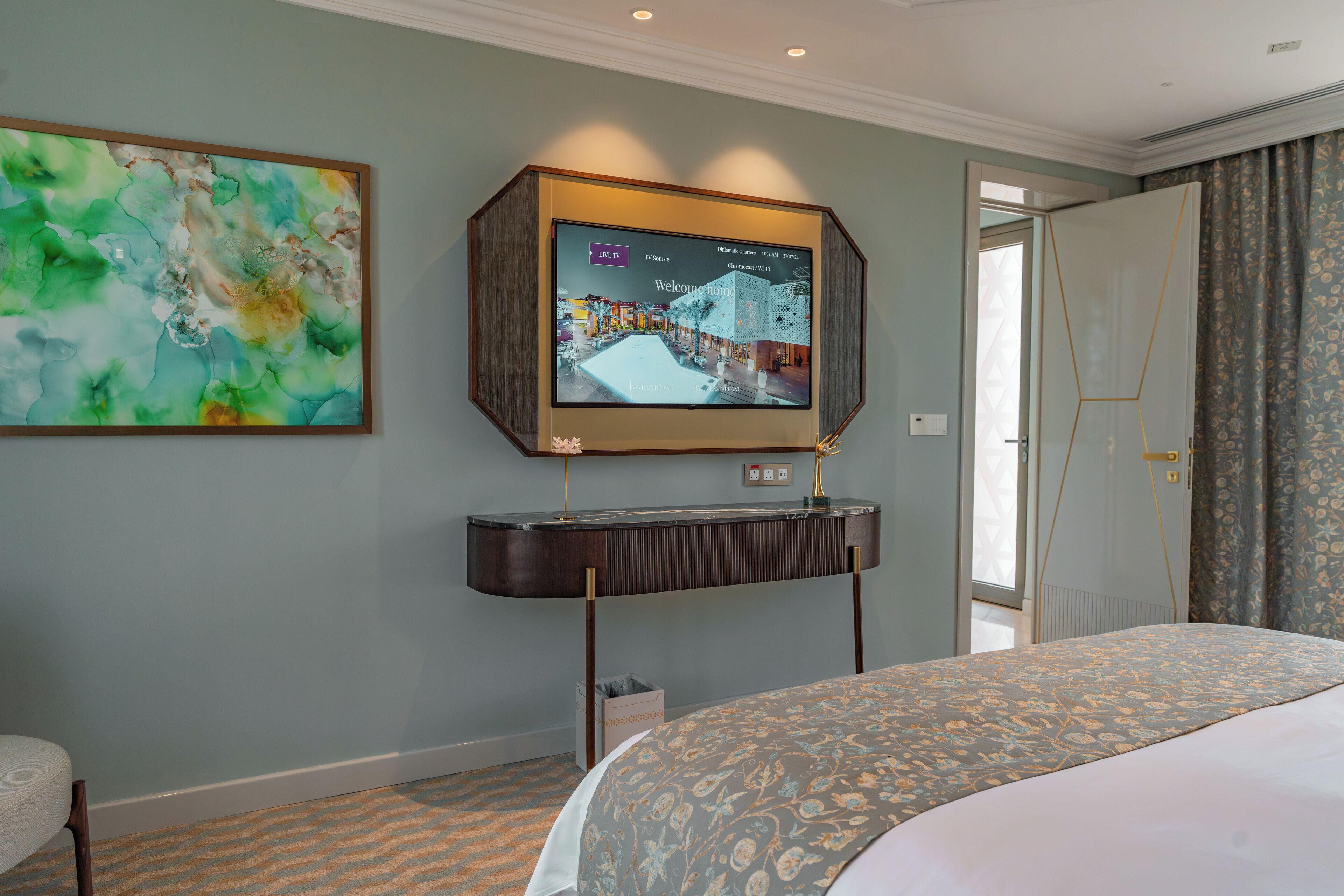 A hotel room with a bed, a flat-screen TV mounted on a wooden console, a large abstract painting, and a door leading to another room.