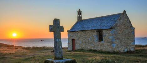 A stone church with a bell tower and a cross in a grassy field at sunset.