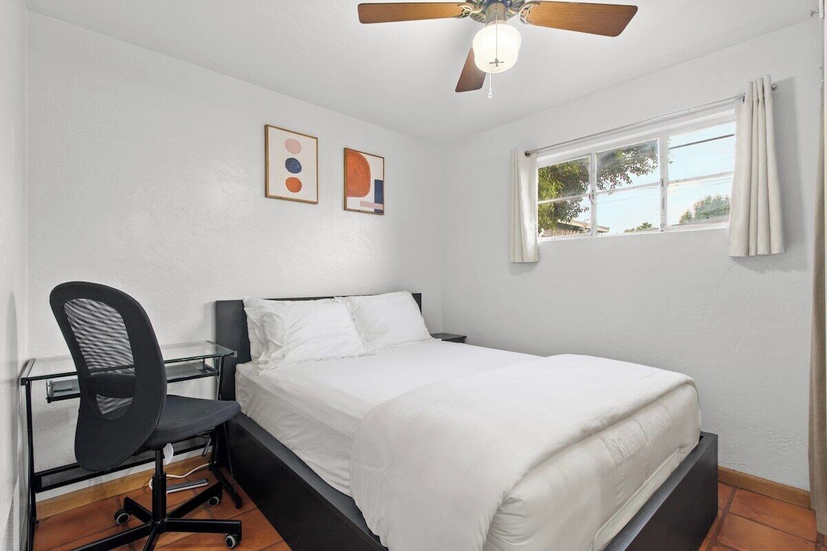 A bedroom with a bed, a desk with a chair, a ceiling fan, and a window with curtains.