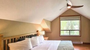Een slaapkamer met een schuine balkenkap, een bed met wit beddengoed, een plafondventilator en een raam met uitzicht op bomen.