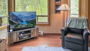 Een woonkamer met een flatscreen televisie op een houten standaard, een leren bank en een groot raam met uitzicht op een bos.