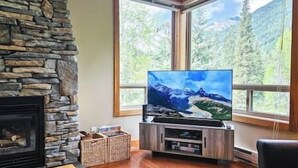 Een woonkamer met een stenen openhaard, een flatscreen televisie op een houten standaard en een groot raam met uitzicht op de bergen.