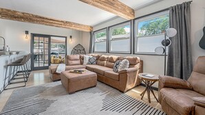 Amplia sala de estar con sofá modular, reposapiés y una mesita redonda. Se aprecia un área de cocina a través de una puerta abierta, y hay ventanas grandes con cortinas.
