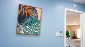A room with a blue wall, a framed tropical leaf artwork, a white door, and a visible kitchen area with white chairs and a table. - Historic Oviedo Charmer - Close to everything (Oviedo)