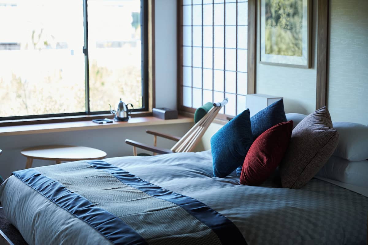 A neatly made bed with a checkered bedspread and pillows, a small round table, a chair, and a window with a view of trees.
