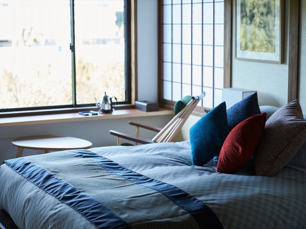 Un dormitorio con cama, almohadas, una ventana y una mesita.