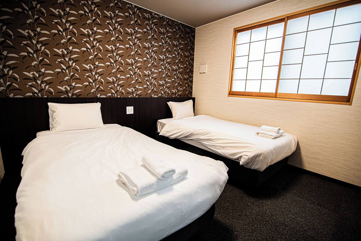 Habitación de hotel con dos camas, papel tapiz estampado, un ventanal grande y suelo alfombrado.