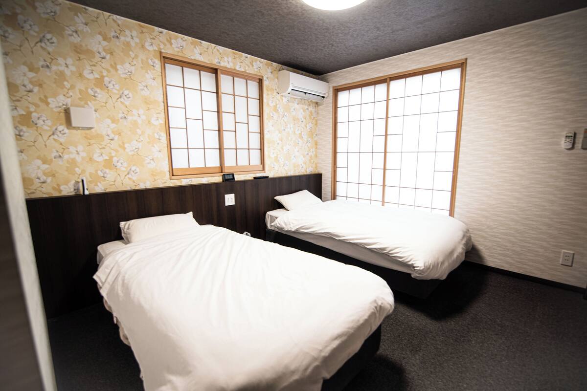 Habitación de hotel con dos camas, papel tapiz floral y ventanas grandes con marcos de madera.