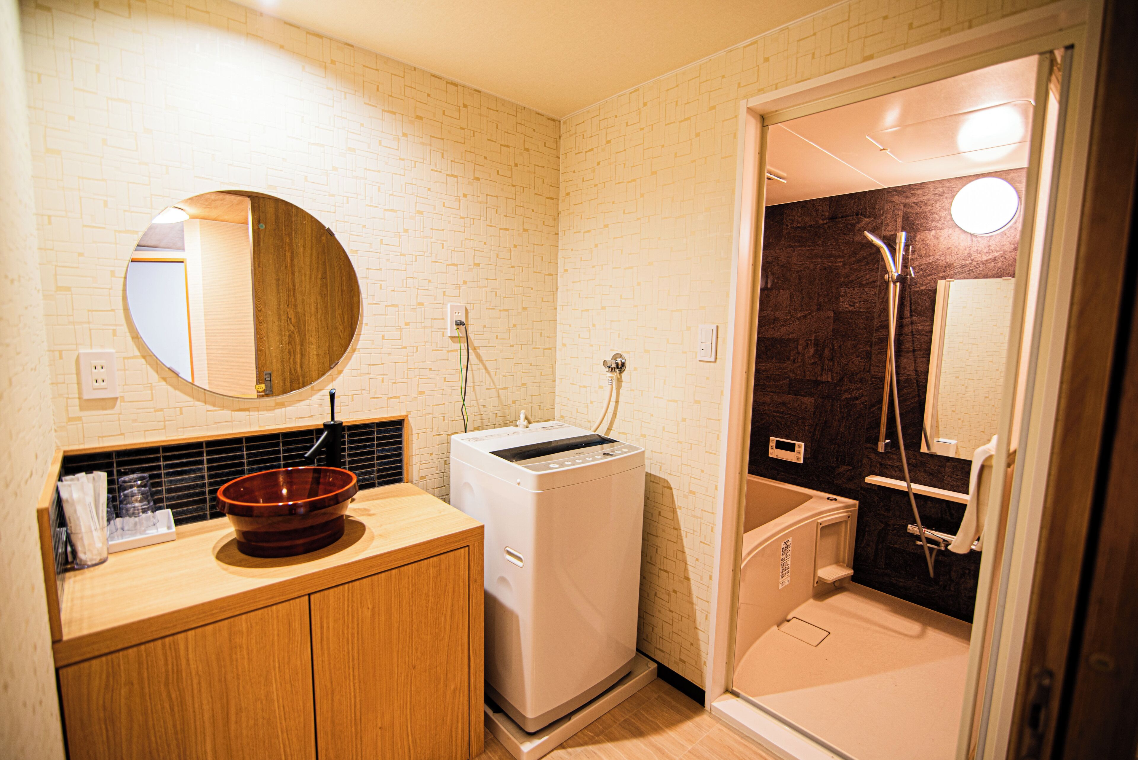 A compact bathroom with a wooden vanity, a round mirror, a washing machine, and a walk-in shower.