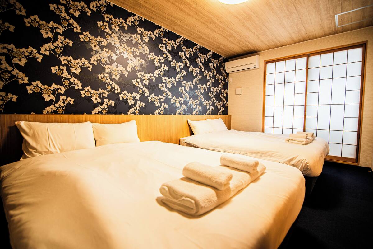 Habitación de hotel con dos camas, papel tapiz floral, un panel corredero tipo shoji y techo de madera.