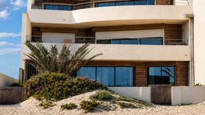Un edificio moderno de varios pisos con balcones, ventanas amplias y un primer plano con arena y plantas.