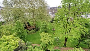 Un jardin luxuriant, avec une variété d’arbres et d’arbustes, un parterre floral circulaire au centre et un banc.