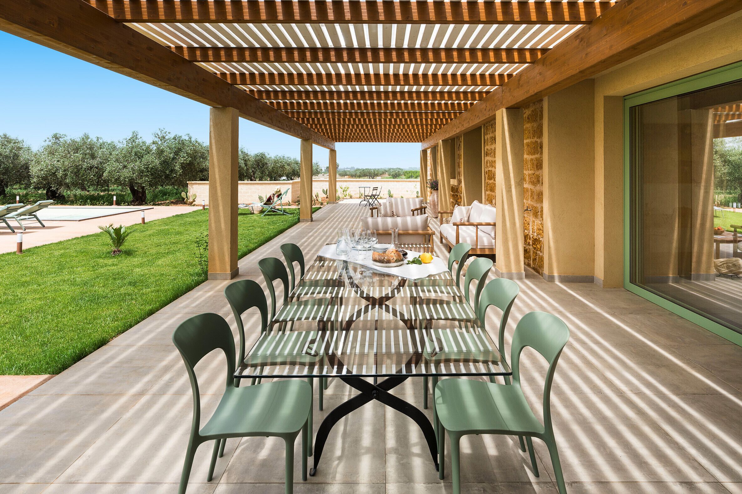 An outdoor patio with a long table and chairs, a pergola overhead, and a view of the surrounding landscape.